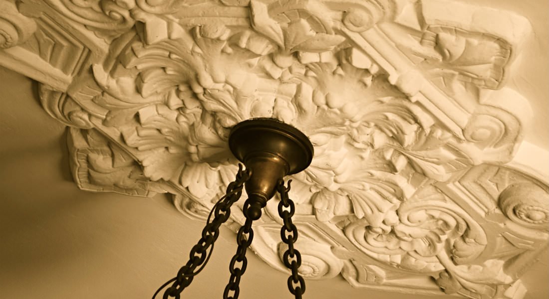 The brass chains and base of a chandilier mount near an ornately patterned decoration on the white ceiling.