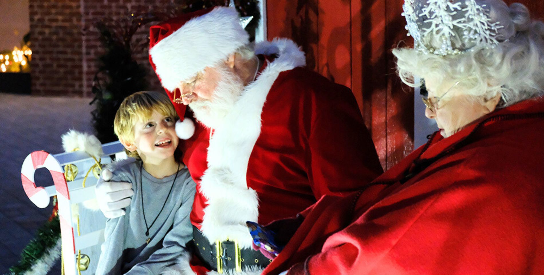 a boy sitting on santa’s lap and smiling up at him with mrs clause sitting next to them