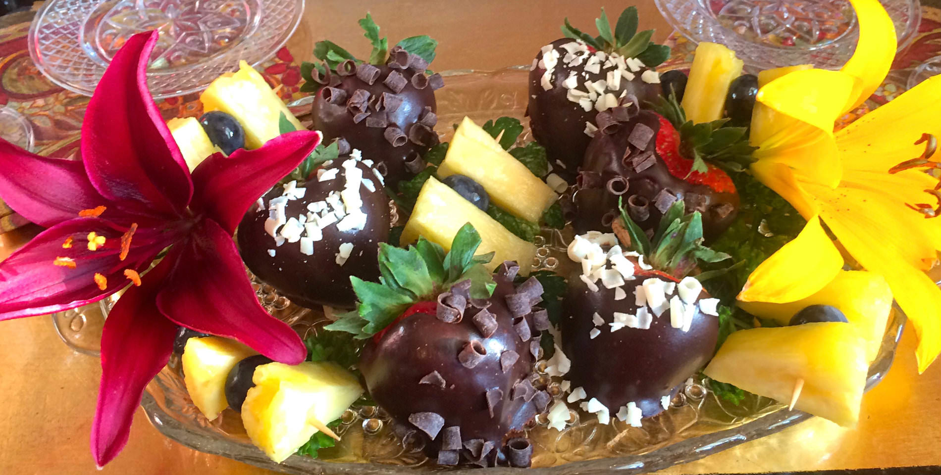 Rich, dark chocolate covered strawberries with green leaves sit in a glass bowl on a table.