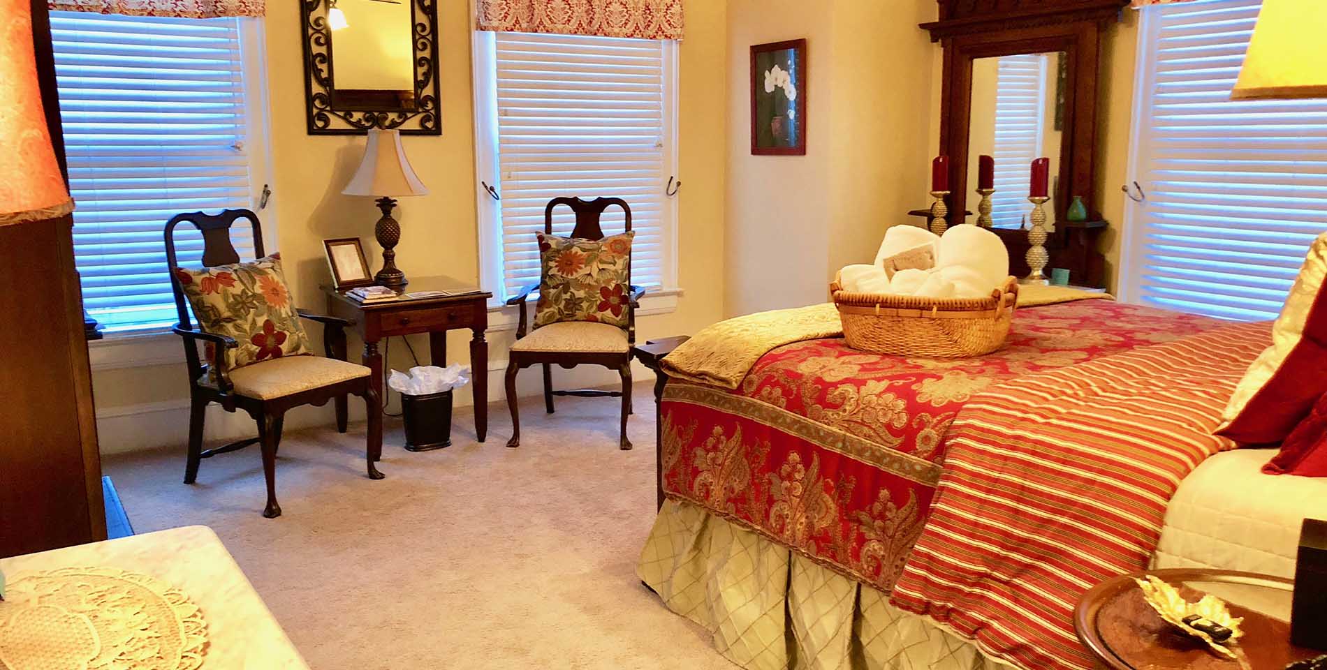 A large bed with red and gold patterned comforter, two chairs and table with floral print pillows atop a beige carpet. 