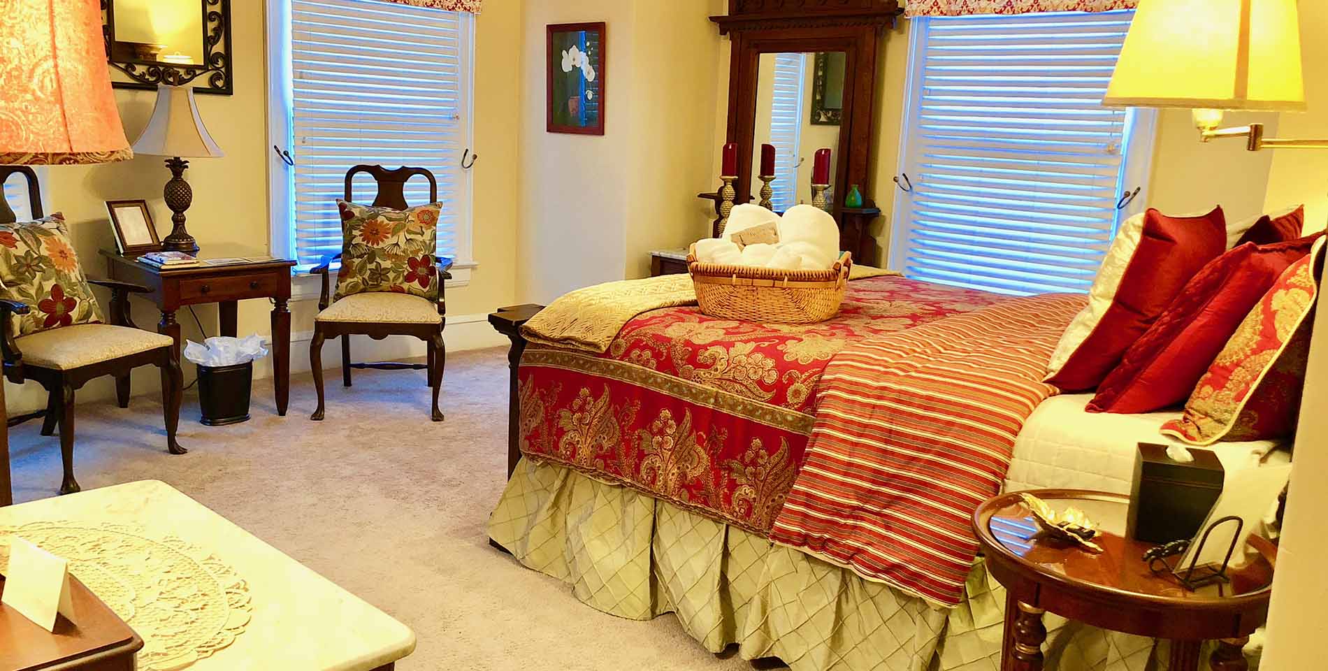 A large bed with a red and gold comforter, red throw pillows and gold bedskirt near a nightstand. 