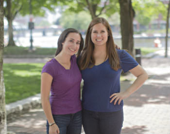 Two women, one with a purple shirt and one with a blue shirt, embrace and smile. 