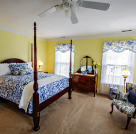 A bed with a blue and white patterned comforter and pillows with beige carpet and yellow walls in the Alyce Wilson room. 