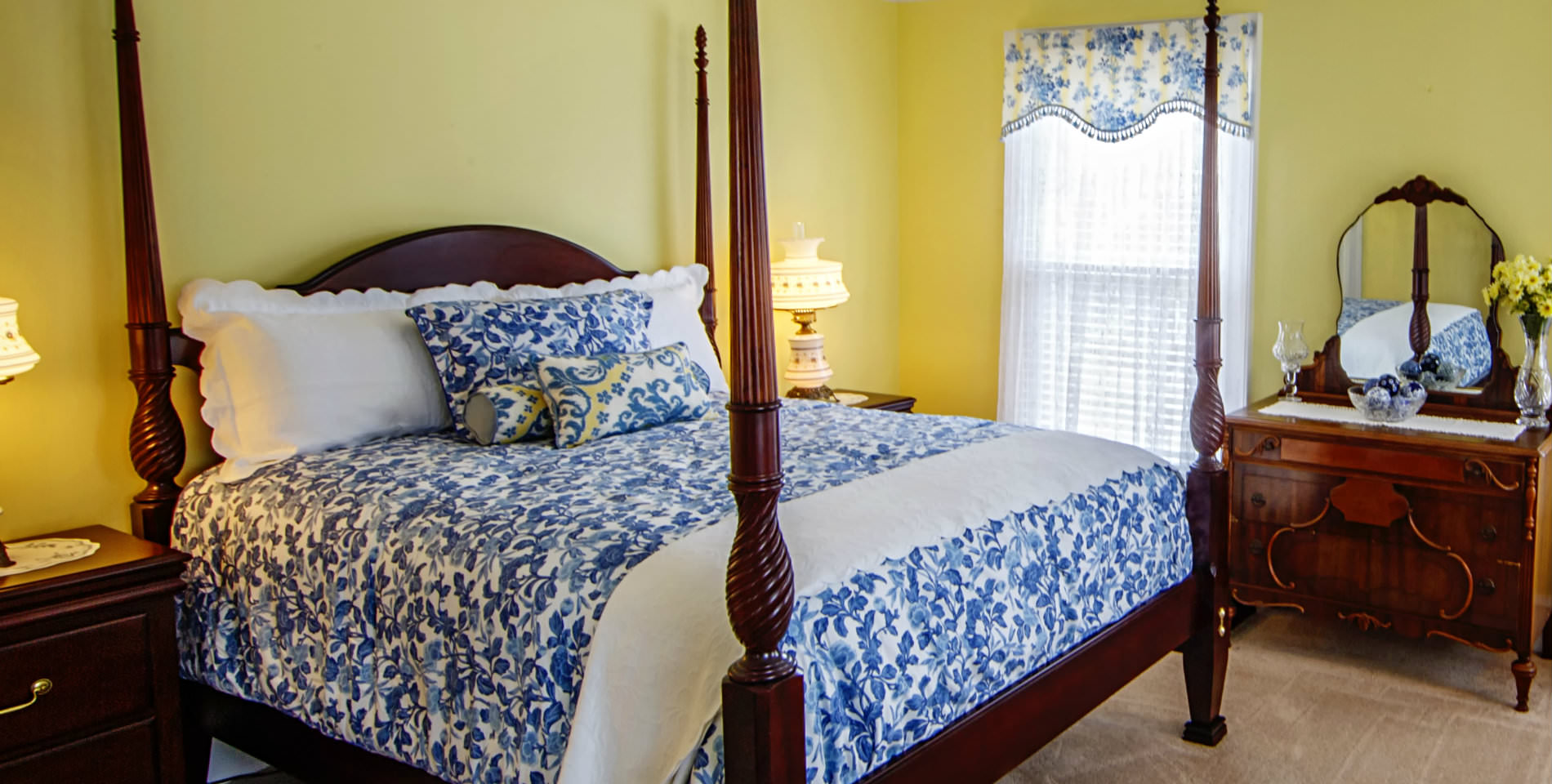 The Alyce Wilson room with a four post wooden bedframe, blue and white bedding and yellow walls. 