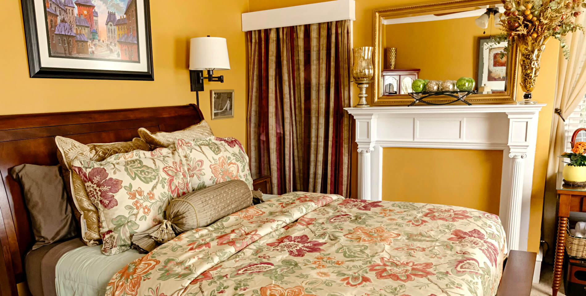 A bed with beige and floral print comforter and pillows and golden yellow walls in the Virginia Wilson room. 