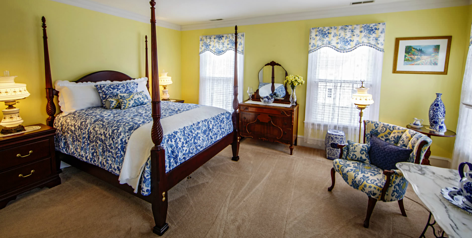 A four post wooden bed, blue and white bedspread, pillows and covered furniture set on a beige carpet with yellow walls. 