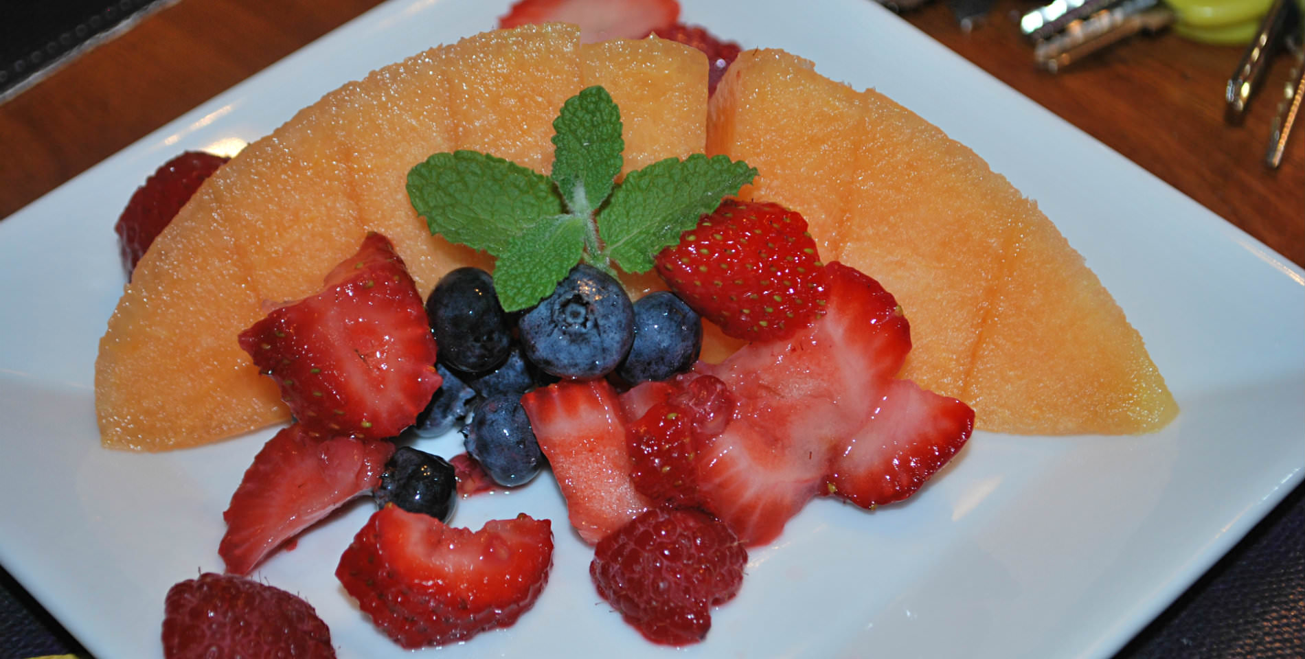 Red strawberries, blueberries and orange cantaloupe served with a sprig of mint on a white plate. 
