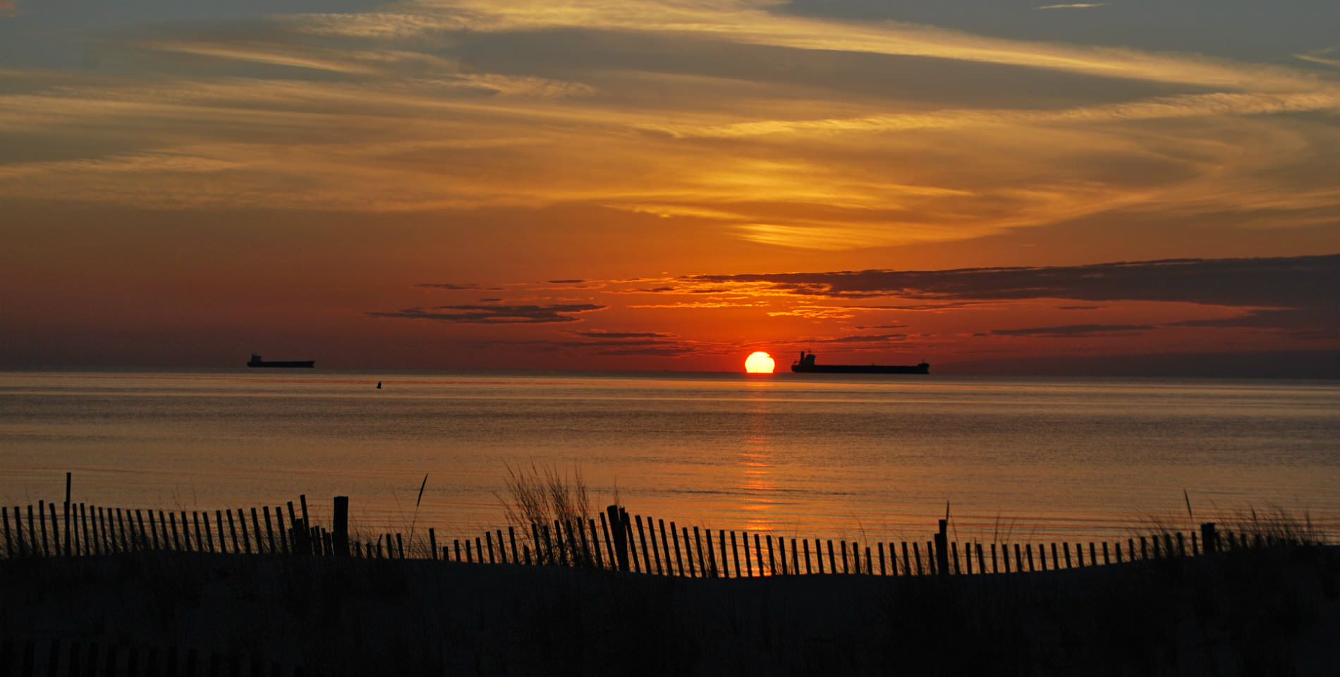A yellow, orange and red sunset over the Bay with a wooden fence lined beach in the foreground. 