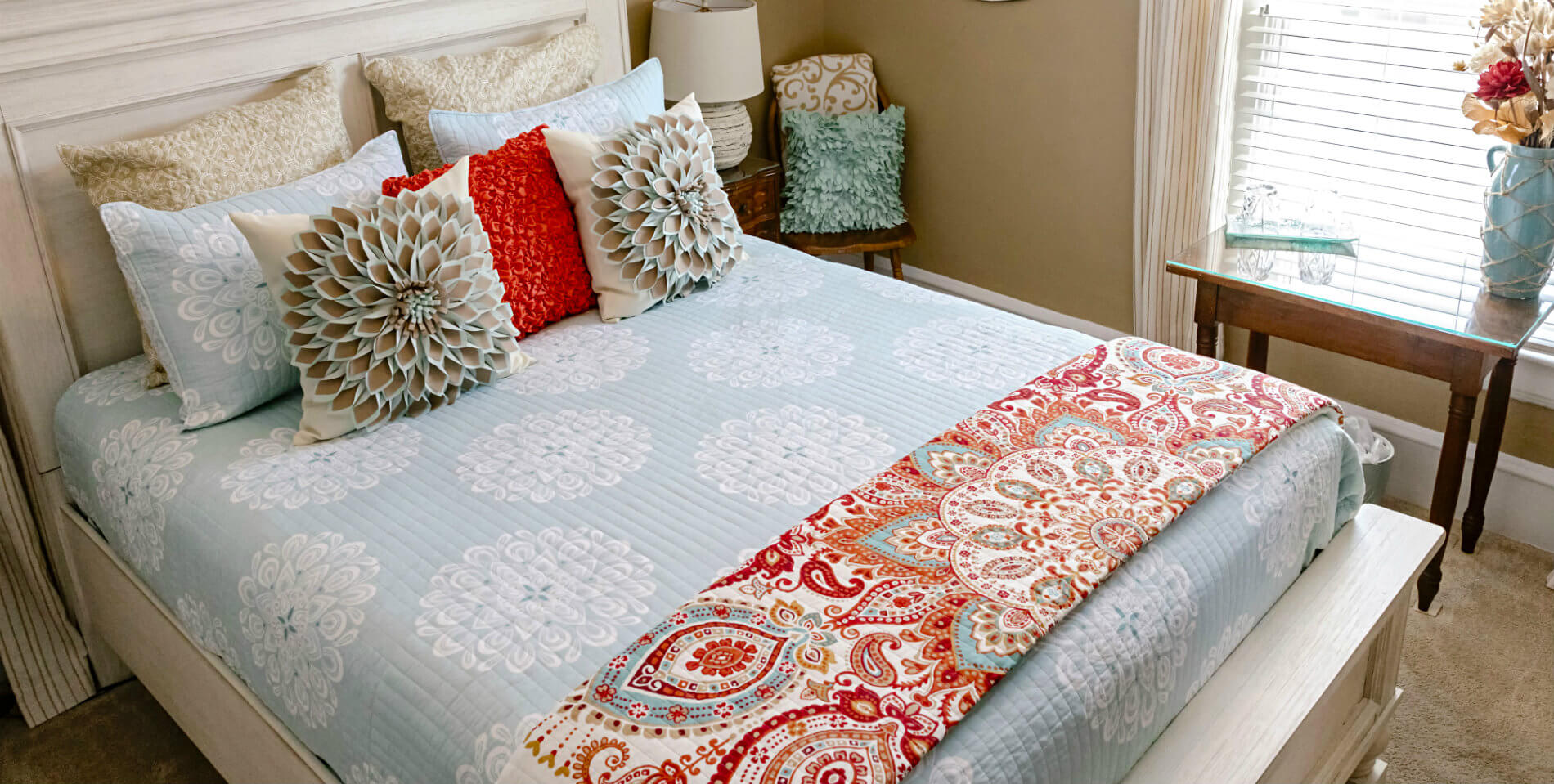 The bed with a light blue and white patterned comforter, red paisley patterned throw and decorative pillows. 