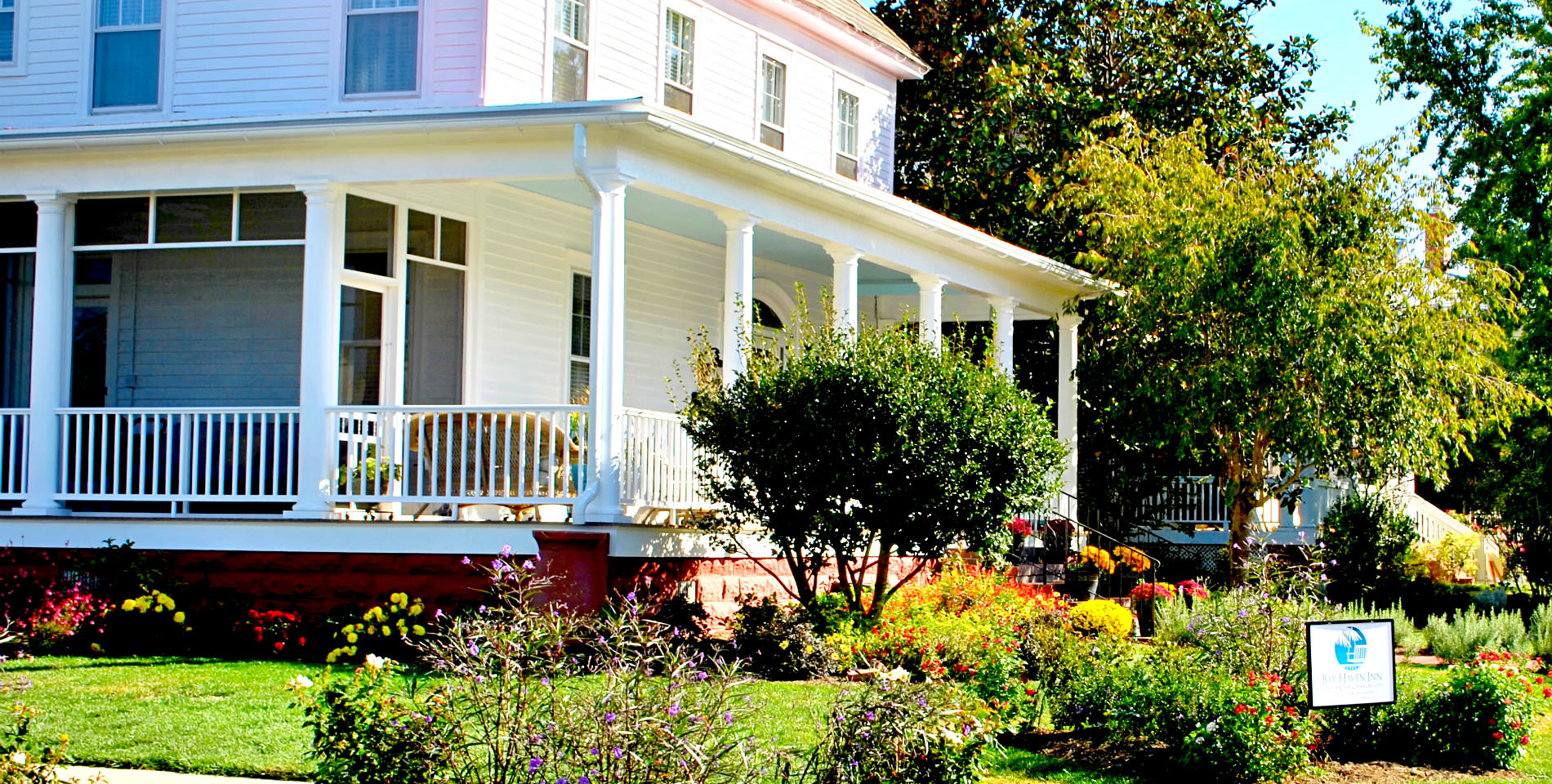 The white siding and wraparound porch of the Inn, with lush green gardens and flowers in the foreground. 