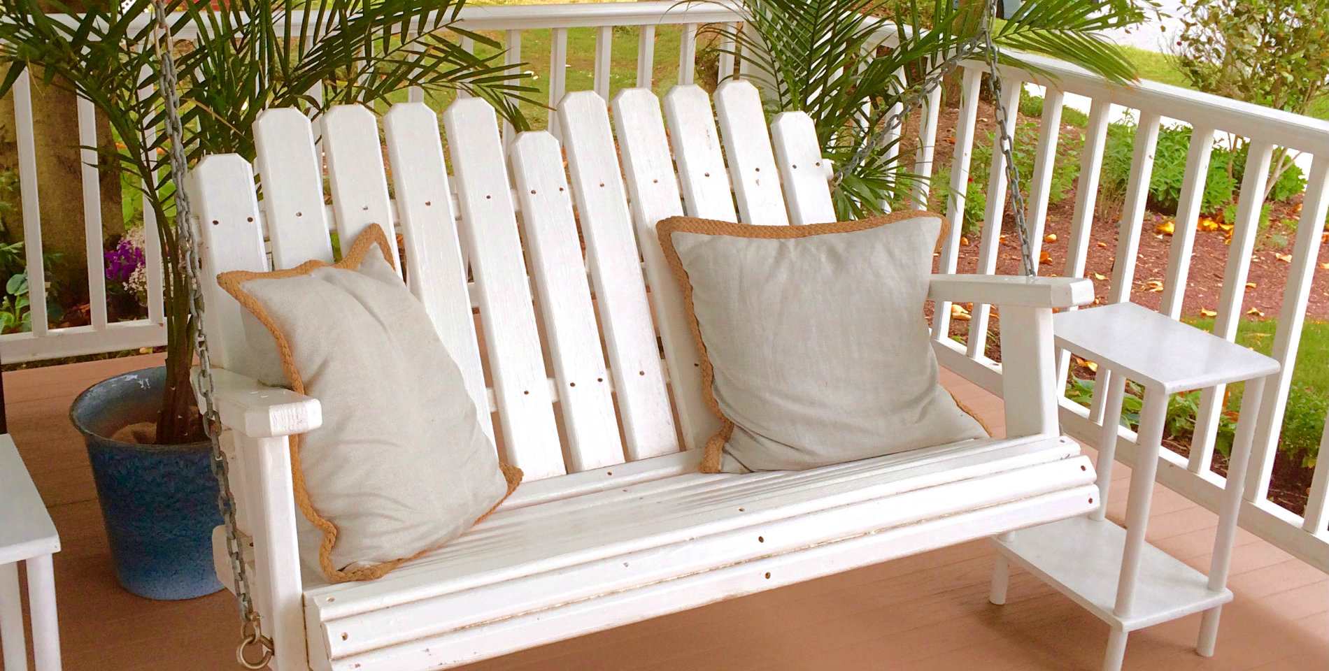A white wooden rocker for two with two beige pillows before two potted palms. 