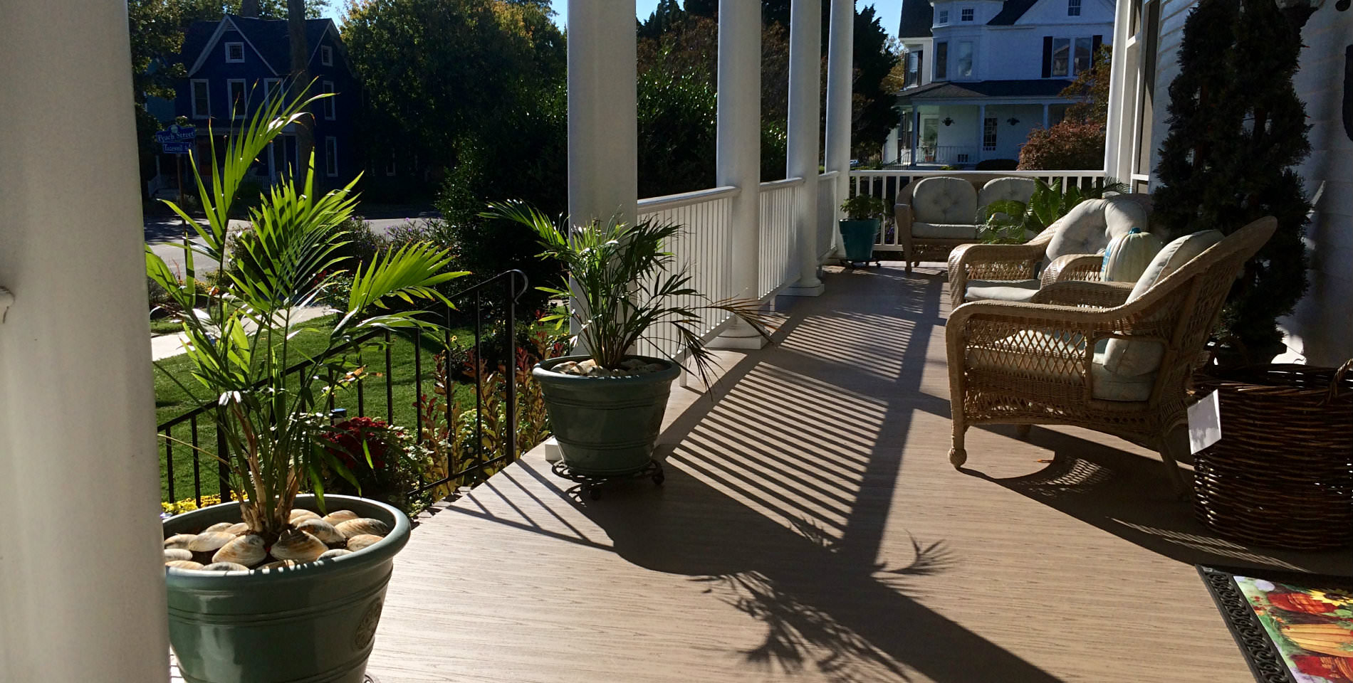 Two potted, green palms and two sitting chairs on the wooden planks of the wraparound porch.  