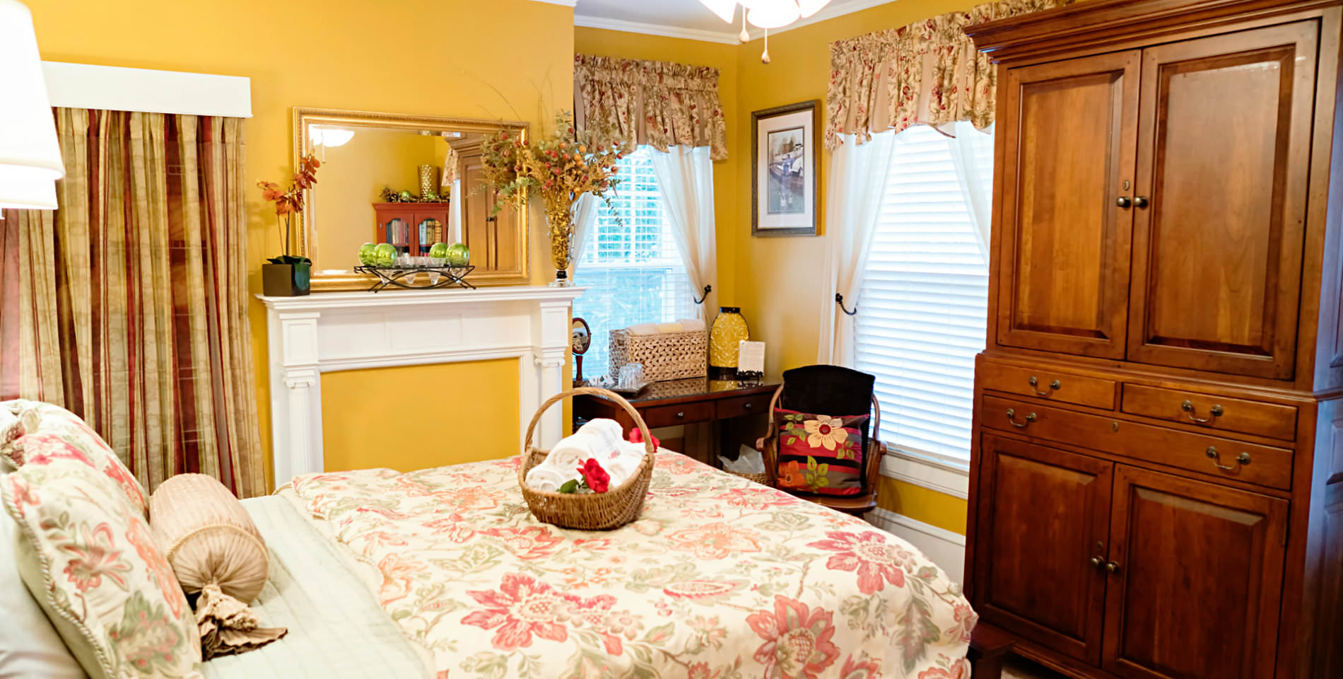 A wood armoire at the foot of a large bed with beige and floral print comforter and throw pillows.