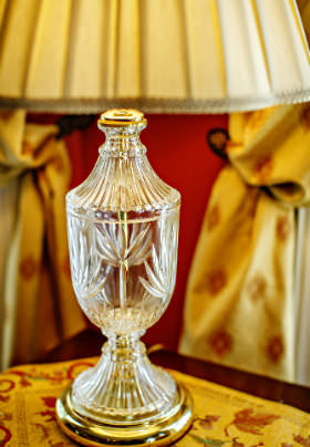 The ornate, glass base of a lamp on a yellow and floral print table cover. 
