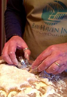 A man carving out chunks of floured dough wearing a beige apron with text: Bay Haven Inn of Cape Charles. 