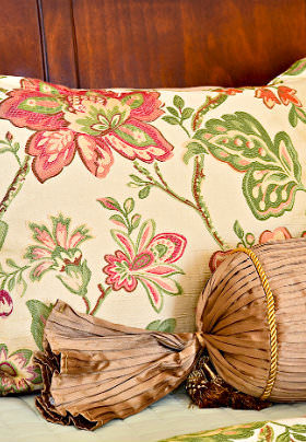 A beige pillow with a red and green floral print before a brown wood paneled wall. 